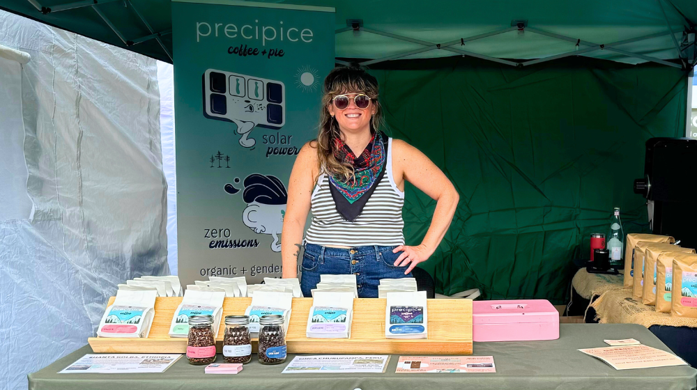 A woman stands in a farmers' market booth with a Precipice Coffee banner behind her and coffee displayed on the table in front of her. The tent is green and encloses the scene.