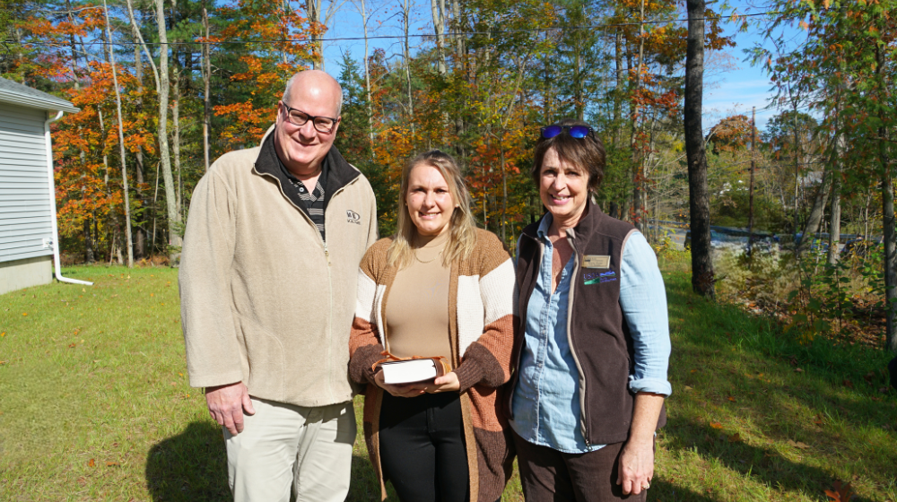 Two USDA staff pose with the homeowner in the grassy front yard of her new home with fall foliage in the background.