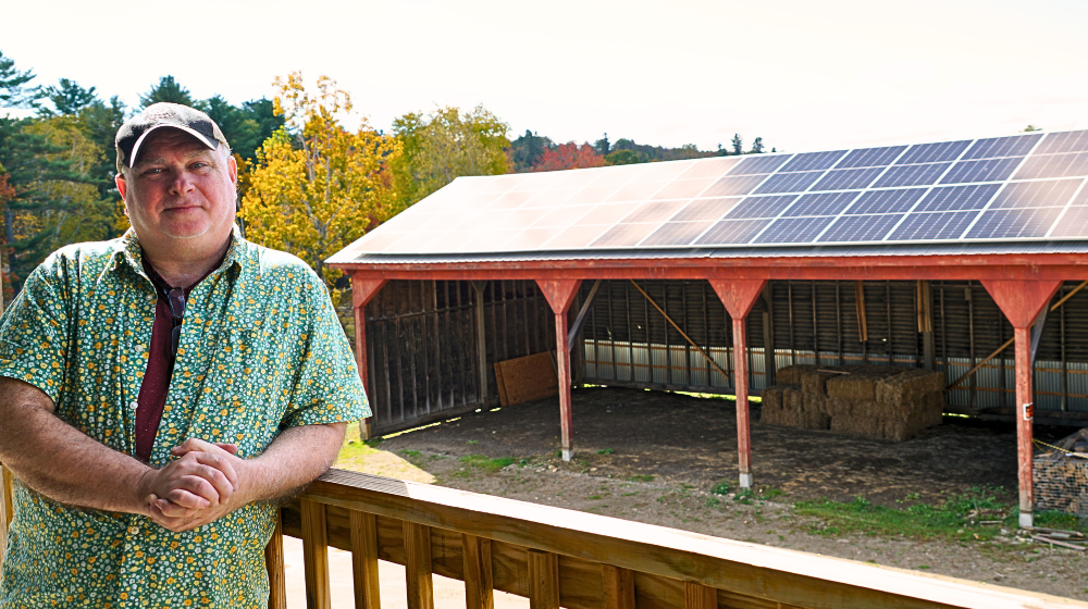 A man stands against a porch railing with a long wood shed behind him. The roof of the shed is covered with solar panels. There is fall foliage in the background.