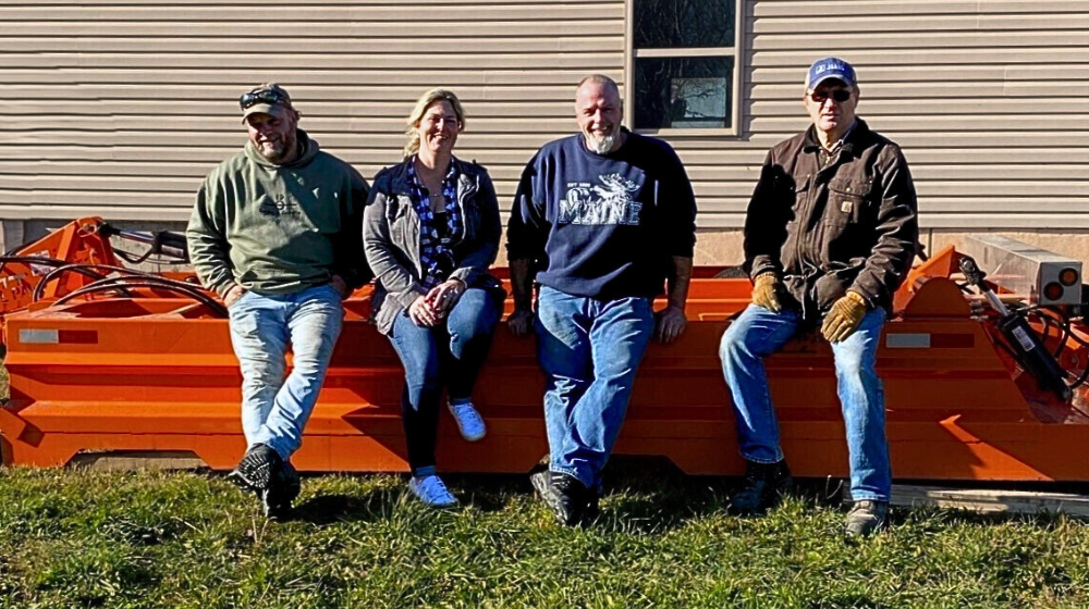 Four members of the Cold Mountain Snowmobile Club are shown seated on an orange snow dragger equipment outdoors under bright sun.