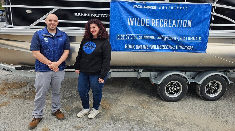 A woman and man stand in front of a pontoon boat with a banner that says the company name, "Wilde Recreation."