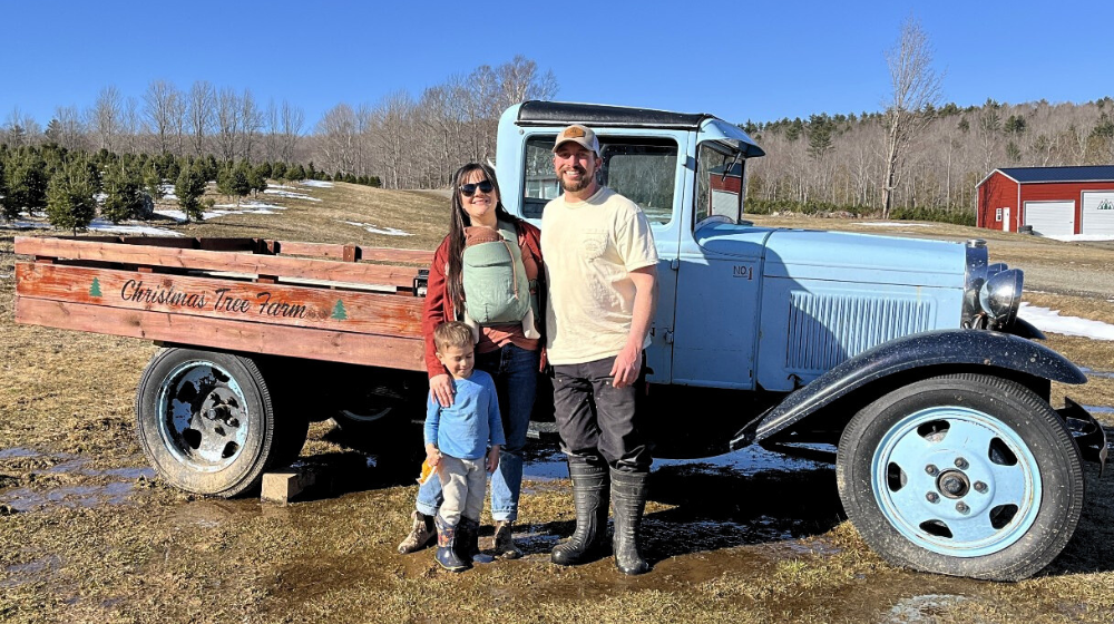 Molly and Jesse of Piper Mountain Christmas Trees stand in front of a vintage blue pickup truck with their two children. In the background there is a red barn and a distant hill.