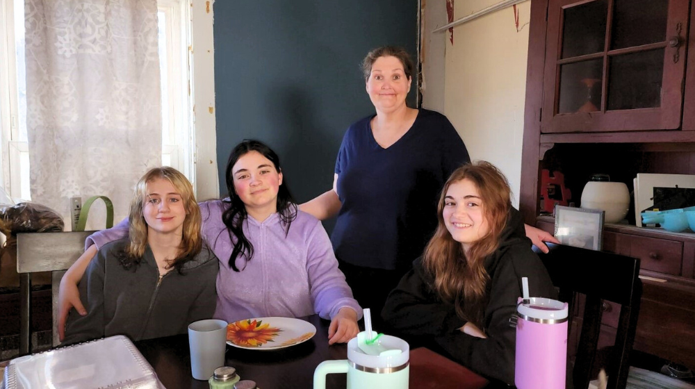 A mother and three daughters pose around a dining room table. There are lace curtains and blue wall in the background and some dishes on the table in the foreground.