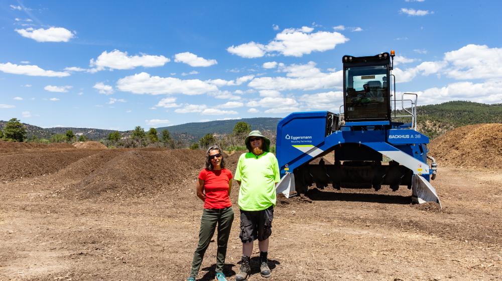 A woman in a red shirt and a man in a green shirt stand in front of a windrow turner machine at an outdoor compost facility. Mounds of compost are behind them under a blue sky with clouds.