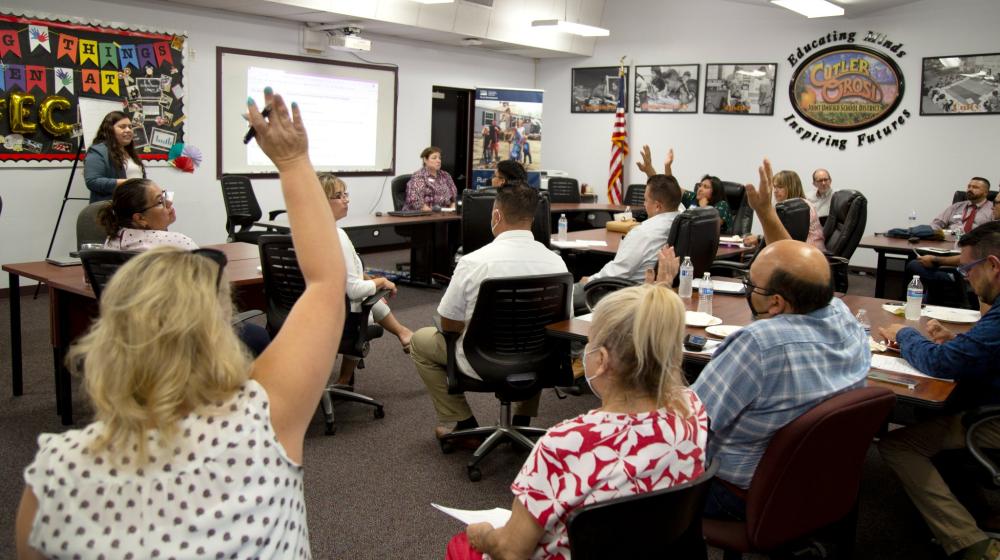 A group of adults sit in a community meeting room, listening to a speaker. Several members of the audience have their hands raised to ask a question.