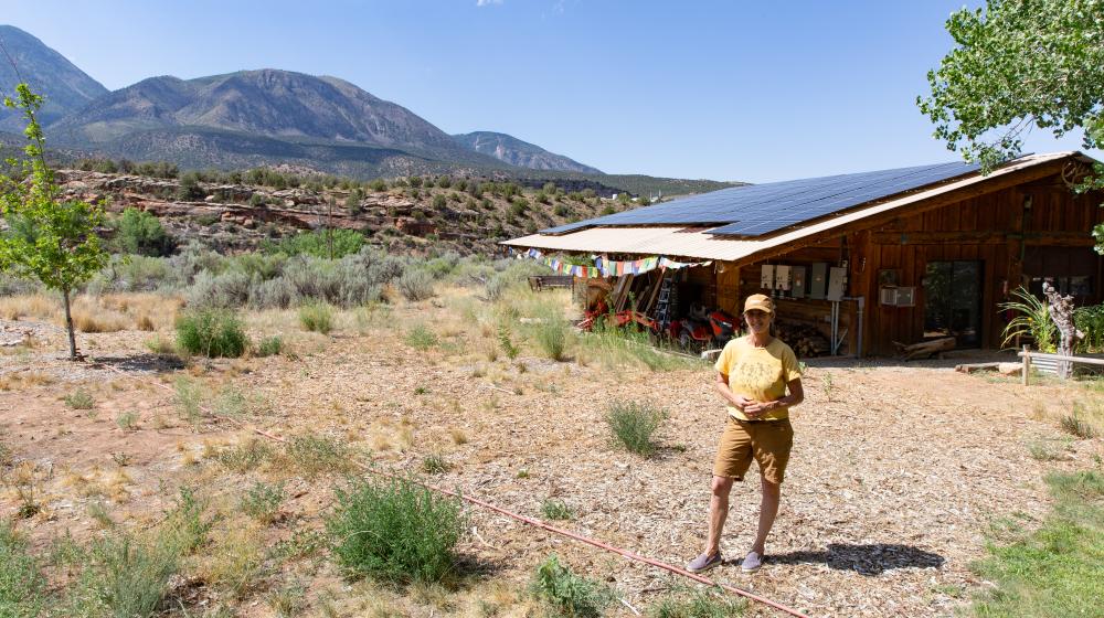 Celine Leavitt stands in front of an outbuilding at her overnight stay guest accommodations in rural Colorado. The building has solar panels mounted on it. In the background is Sleeping Ute Mountain.