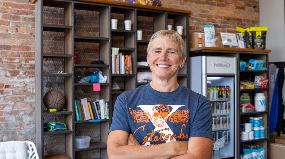 A woman stands in the lobby area of a small gym in rural Colorado. A teal colored sign with a skull and wing logo for Catacombs Fitness Collective is displayed on the wall behind her.
