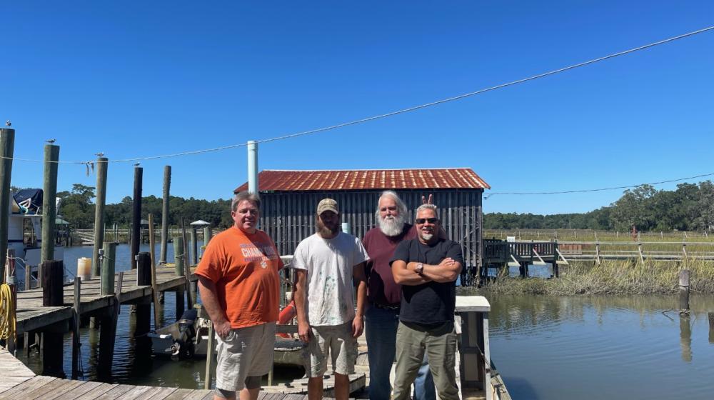 Four smiling men stand together on a dock next to the water. There is a building on the shore in the background.