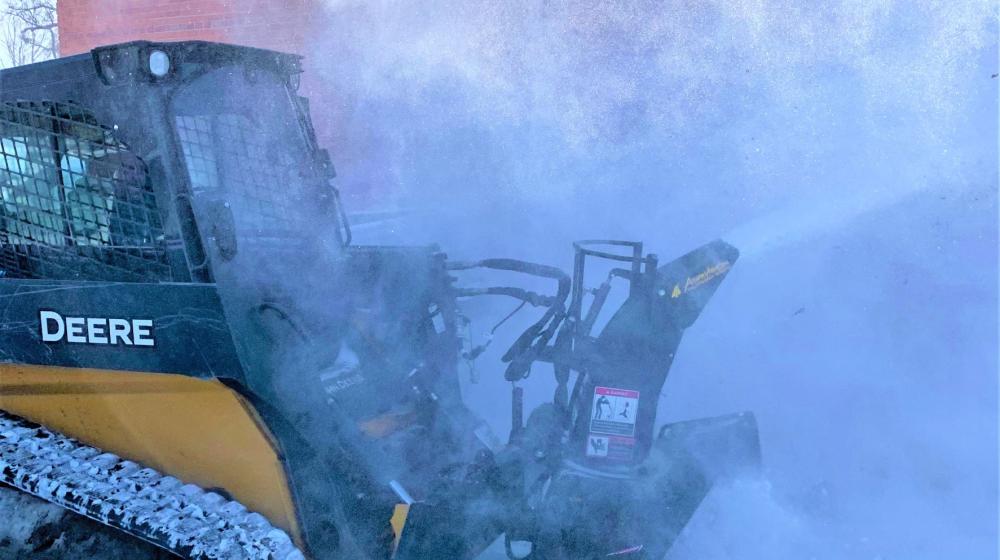 A skid steer with snow blower attachment blowing snow off a sidewalk