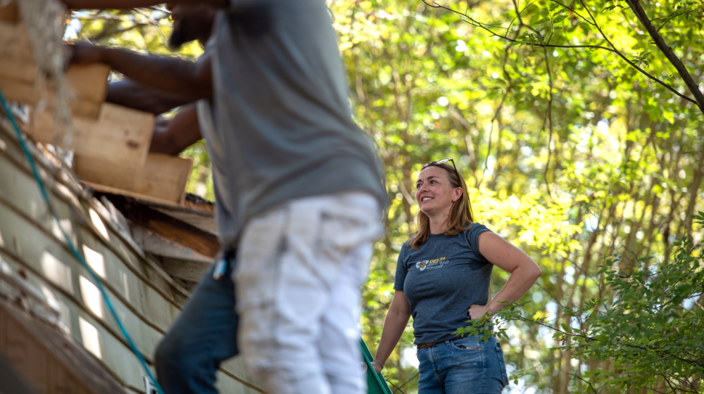 A smiling woman stands on the roof of a house and watches while works fix the roof.