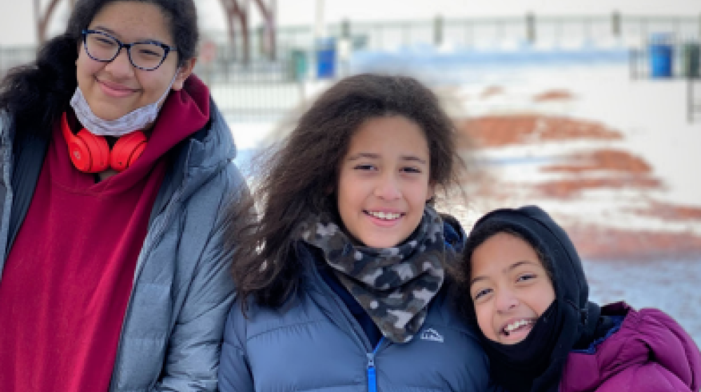Three smiling girls in winter coats in front of a snow covered field.