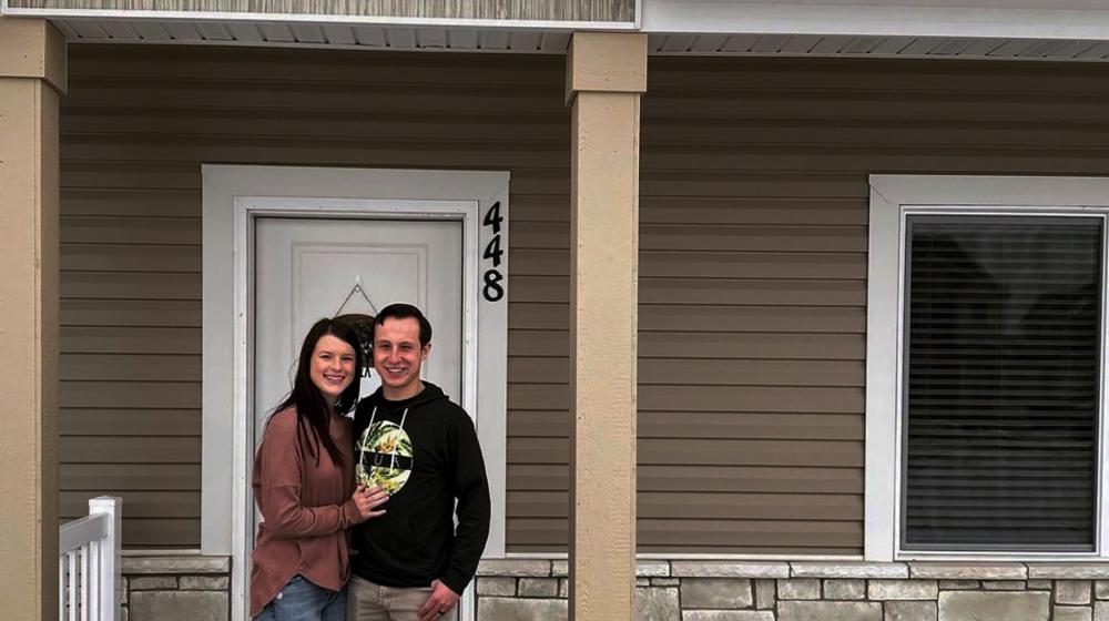 Young Caucasian couple with dark colored hair and clothing stand in front of a home with brown siding and tan brick.