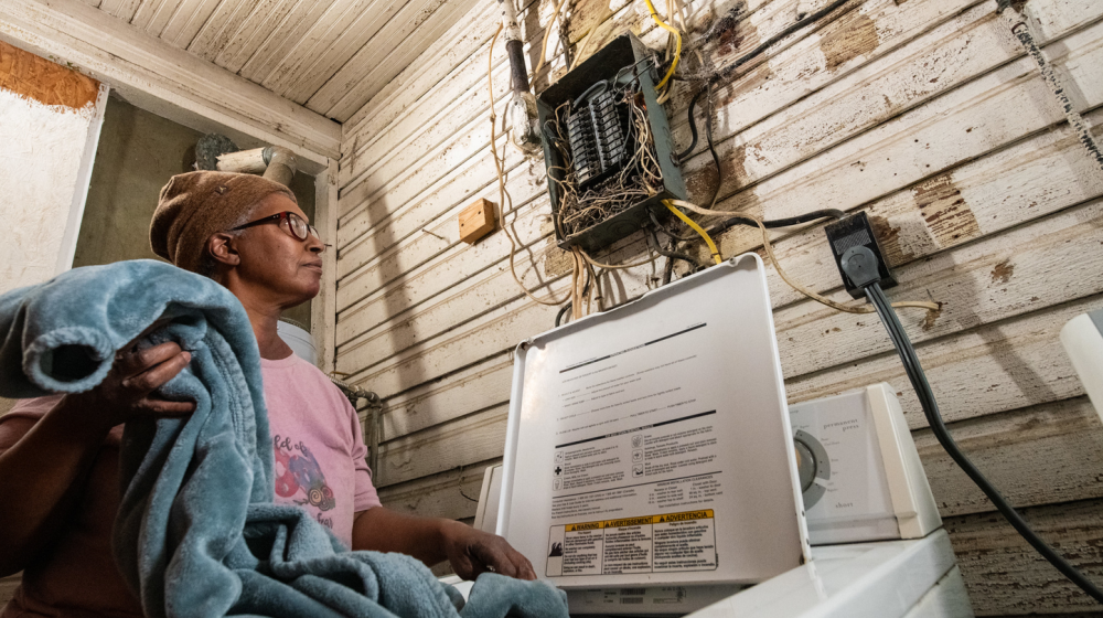 Women putting a blanket in the washing machine while looking at an electrical box above the washer that has no cover and is a nest of wires leading into the fuses. It looks very unsafe.