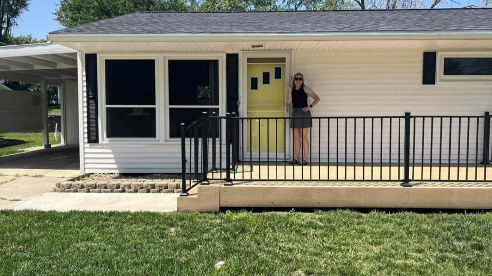 A woman stands in front of a white house with a metal railing