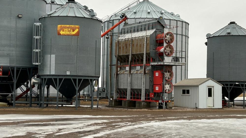 A photo on a farm with a grain dryer in the center. There is a small white building to the right and grain bins around the grain dryer.