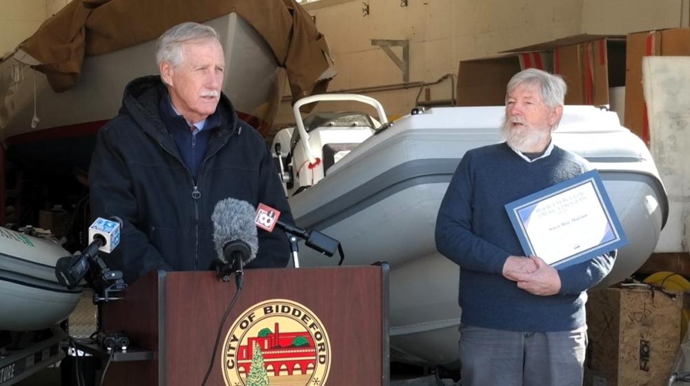 Senator Angus King stands at a podium bearing the seal of the City of Biddeford. Sean Tarpey stands to the right in the photo, holding an award signifying Saco Bay Marine's REAP grant. In the background are 2 electric boats and a larger boat in the far background.