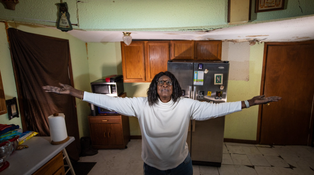 A women standing in her house with her arms held out at shoulder height to show how dipped her roof is. It sags in the middle where she stands.