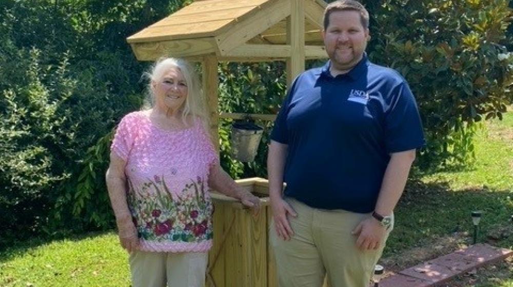 Smiling elderly woman next to a younger man standing in front of a wooden structure that covers a well in a green lawn.