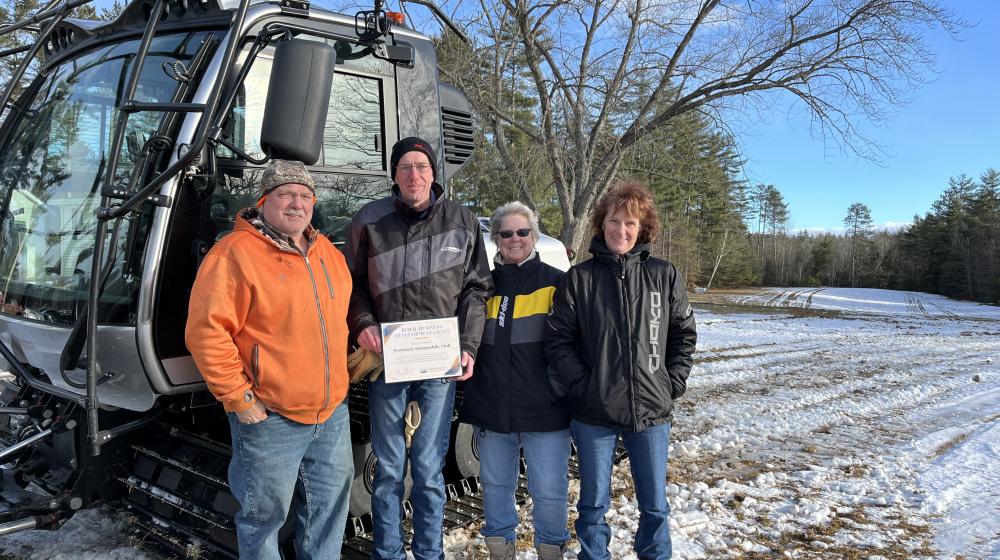 Two men and two women stand in front of a snow grooming machine in a snowy field under a blue sky. They are dressed warmly, and there are leafless trees in the background.