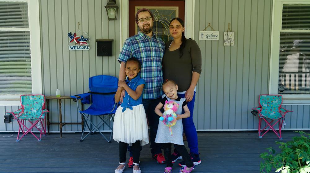 A smiling family, father, mother, two young daughters, standing on the porch of their own home.