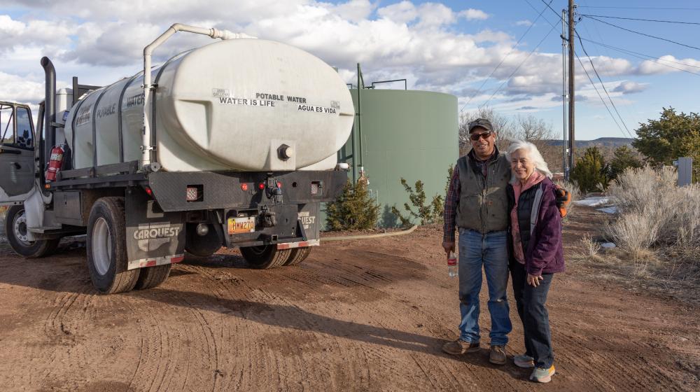 Two people standing in front of a water tank and water truck