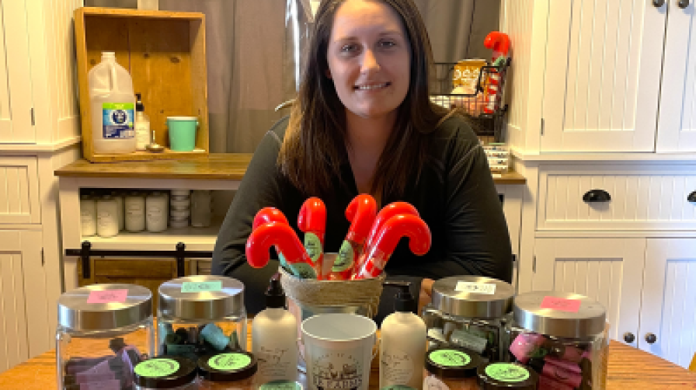 Woman sitting at table with home made soaps, lotions, and balms.