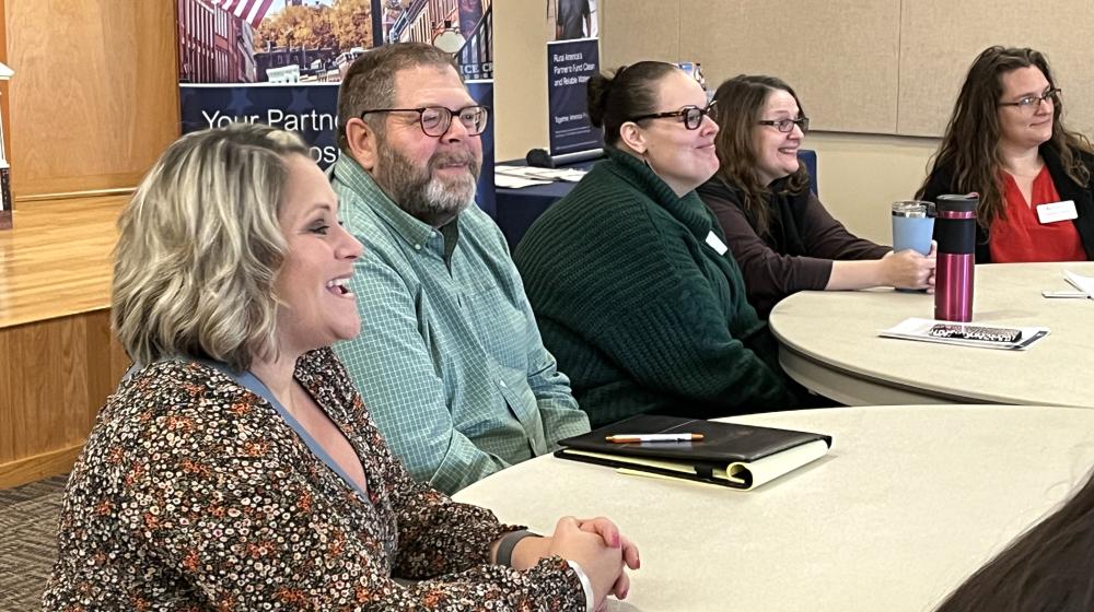 Four women and one man sit at tables in a large room with a USDA banner in the background.