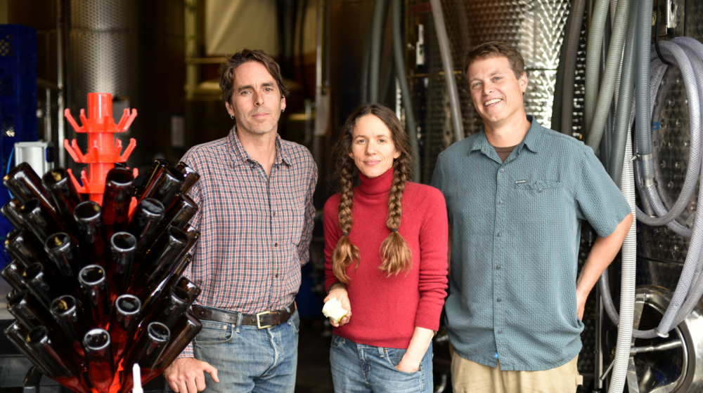 finnriver coowners photo by james curtis. Three people smiling in front of fermenting equipment and bottles.