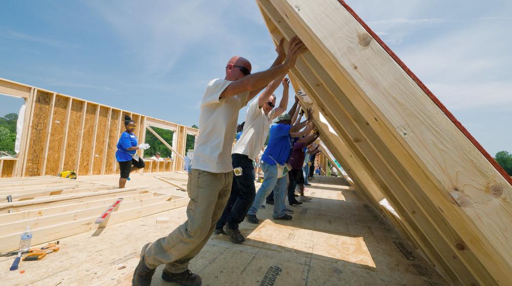 Photo of people lifting up a wall frame they have built out of wood while building a house.