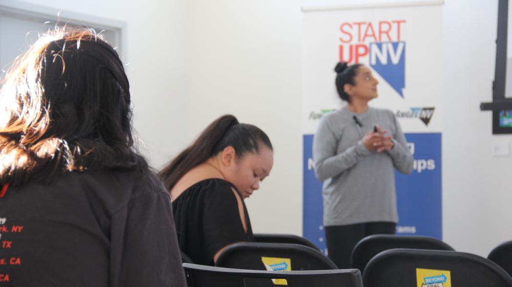 picture of presenter looking off frame towards a projector screen. behind the presenter is a banner that says startup nv.