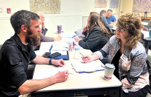 A man and a woman are pictured seated at a table, talking and taking notes. Other people work at the same table and other tables in the background. There is abstract artwork on the walls in the background.