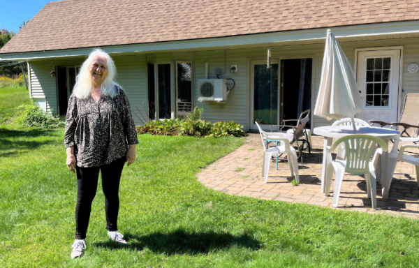 A woman stands before a cream colored house set into a hillside, with a brick patio and white patio furniture arranged. There is a green lawn in the foreground and trees in the background in bright sunshine.