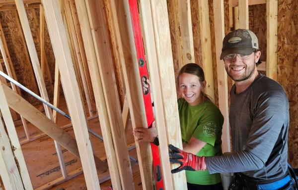 Cody (Caucasian white male wearing black and grey jacket with matching ballcap) and Melissa (Caucasian young adult female wearing olive green shirt)Olsen in Hyrum, Utah in June of 2024, seen standing within the wooden framework of their Mutual Self-Help house.