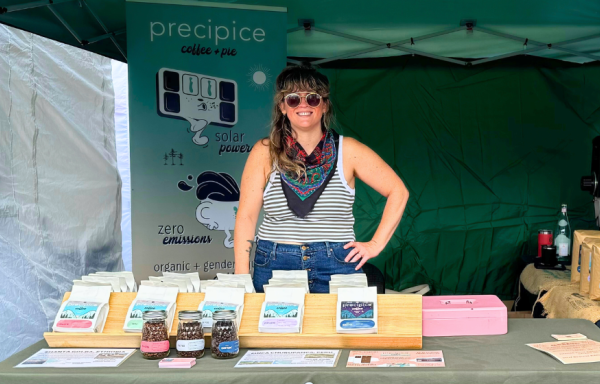 A woman stands in a farmers' market booth with a Precipice Coffee banner behind her and coffee displayed on the table in front of her. The tent is green and encloses the scene. 