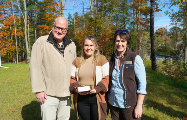 Two USDA staff pose with the homeowner in the grassy front yard of her new home with fall foliage in the background.
