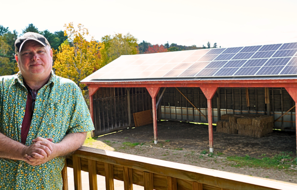 A man stands against a porch railing with a long wood shed behind him. The roof of the shed is covered with solar panels. There is fall foliage in the background.