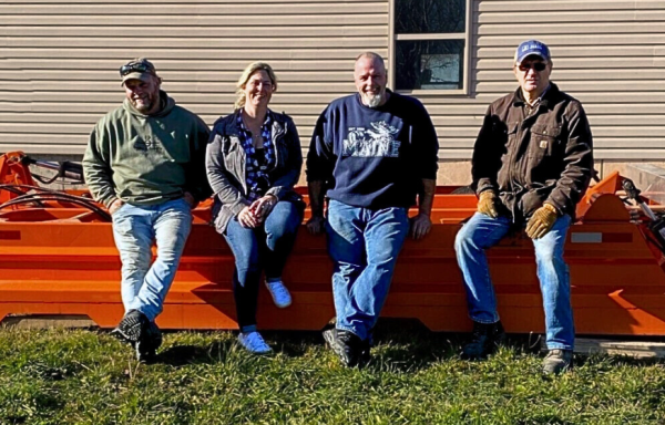 Four members of the Cold Mountain Snowmobile Club are shown seated on an orange snow dragger equipment outdoors under bright sun.