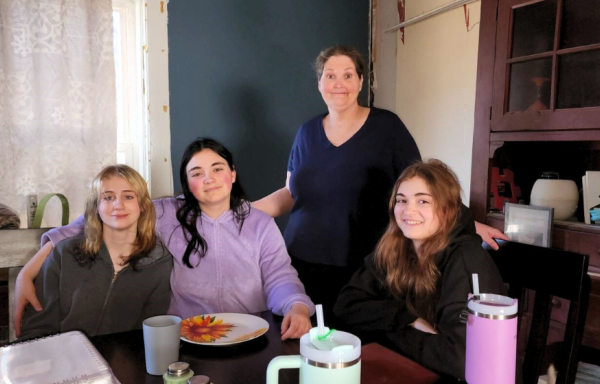 A mother and three daughters pose around a dining room table. There are lace curtains and blue wall in the background and some dishes on the table in the foreground.