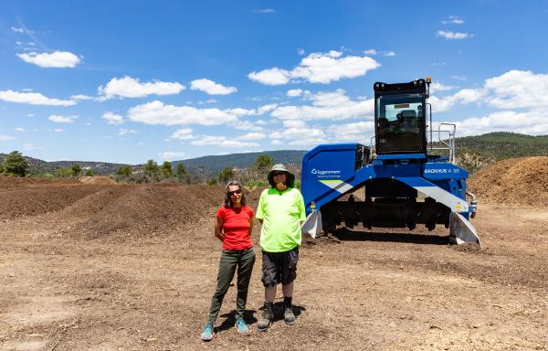 A woman in a red shirt and a man in a green shirt stand in front of a windrow turner machine at an outdoor compost facility. Mounds of compost are behind them under a blue sky with clouds.