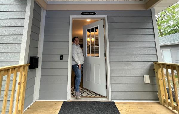 Smiling woman standing in the open doorway of her home as seen from the porch of the home.
