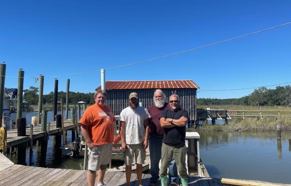 Four smiling men stand together on a dock next to the water. There is a building on the shore in the background.