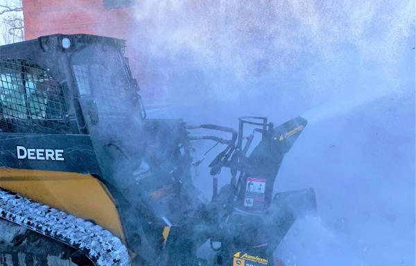 A skid steer with snow blower attachment blowing snow off a sidewalk