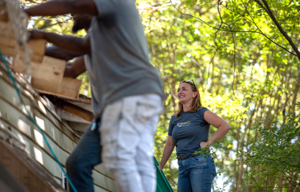 A smiling woman stands on the roof of a house and watches while works fix the roof.