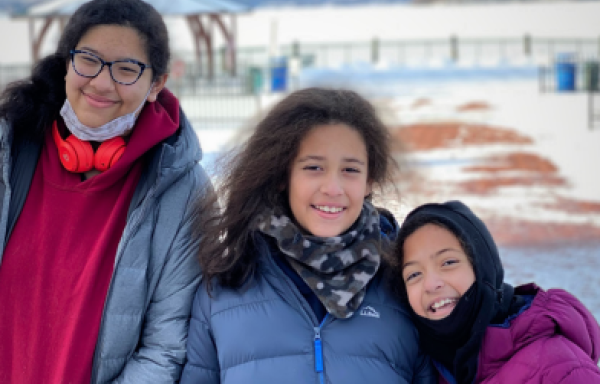 Three smiling girls in winter coats in front of a snow covered field.