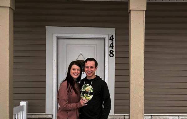 Caucasian couple with light skin, dark hair, stand on porch of their new house with brown siding and tan brick.