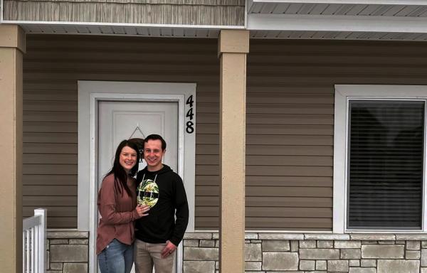 Young Caucasian couple with dark colored hair and clothing stand in front of a home with brown siding and tan brick.