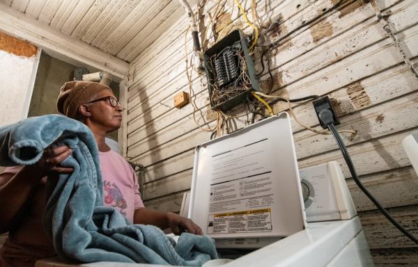 Women putting a blanket in the washing machine while looking at an electrical box above the washer that has no cover and is a nest of wires leading into the fuses. It looks very unsafe.