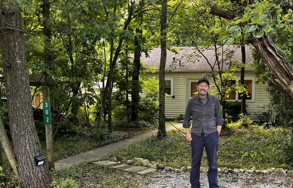 Man standing in front of house on wooded lot. 