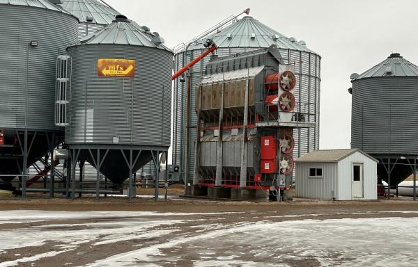A photo on a farm with a grain dryer in the center. There is a small white building to the right and grain bins around the grain dryer.