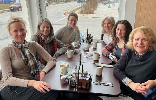 Six women sit around a table drinking coffee and tea